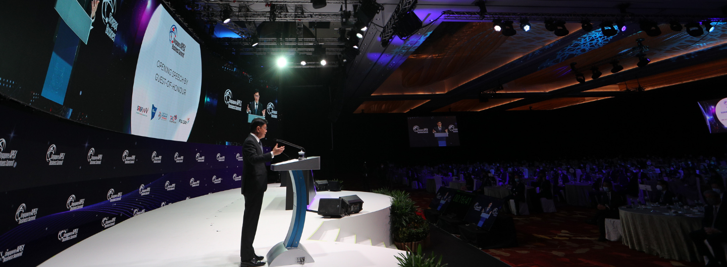 Speaker at podium on stage at Singapore Apex Business Summit, audience seated. Large screens display event logos.